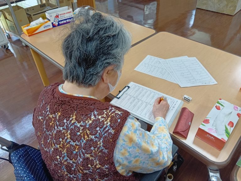 高齢者が食堂のテーブルで書類に記入している様子。レクリエーション活動と思われる。